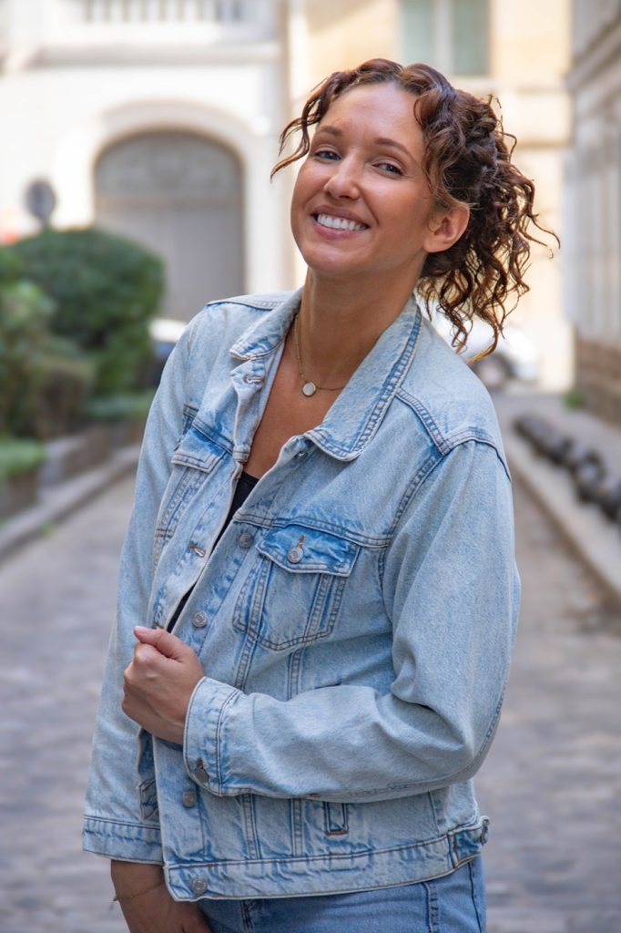 Cora Tellaa souriante, veste en jean, dans une rue pavée